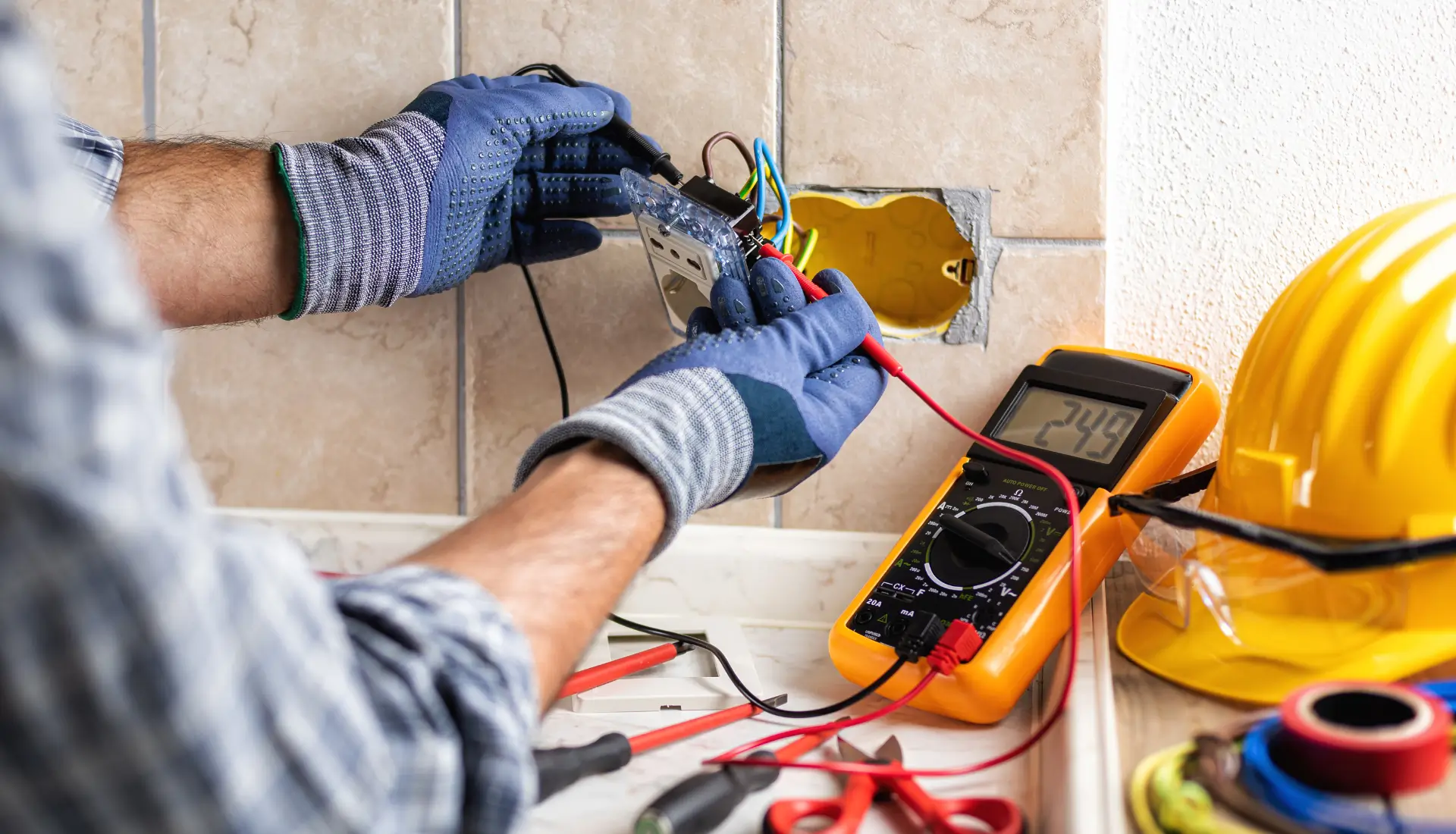 A close-up of an electrician wearing gloves, working with exposed wires and a socket inside a wall. A digital multimeter displays a voltage reading, with various tools and a yellow hard hat nearby.