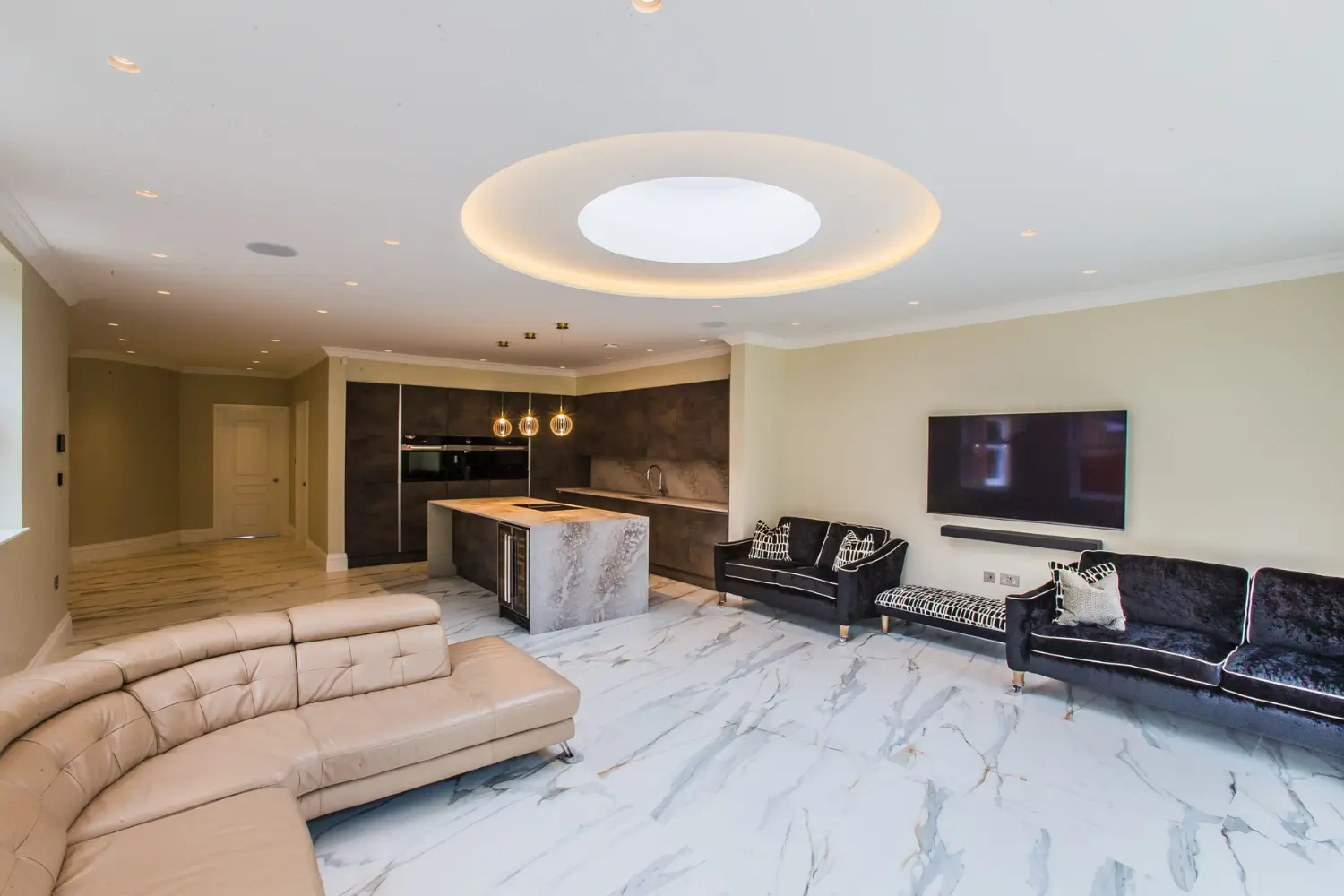 Modern open-plan kitchen and living room featuring a circular ceiling light, marble flooring, beige and black sofas, and a mounted television, designed by Rajvar Technologies.