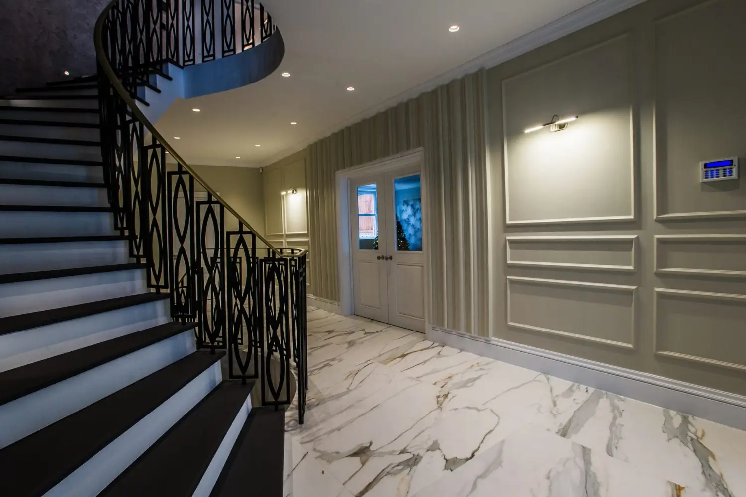Elegant hallway with a sweeping black staircase featuring decorative railings, marble flooring, panelled walls, and soft recessed lighting, designed by Rajvar Technologies.