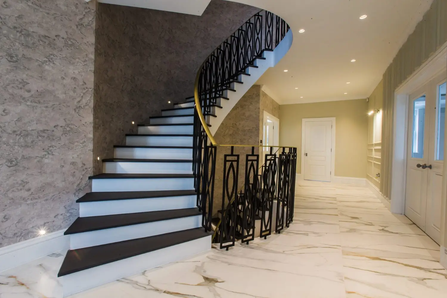Elegant hallway with a sweeping black staircase featuring decorative railings, marble flooring, panelled walls, and soft recessed lighting, designed by Rajvar Technologies.