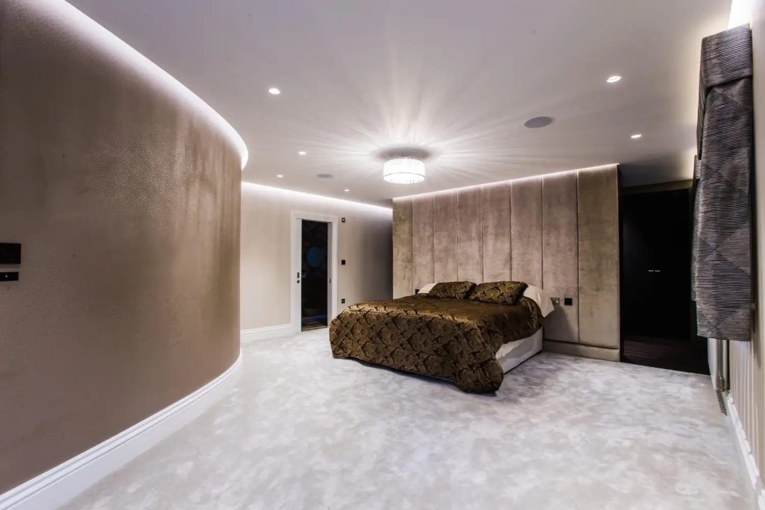 Modern bedroom with a curved feature wall, plush carpet, upholstered headboard wall, ambient LED lighting, and a central chandelier, designed by Rajvar Technologies.
