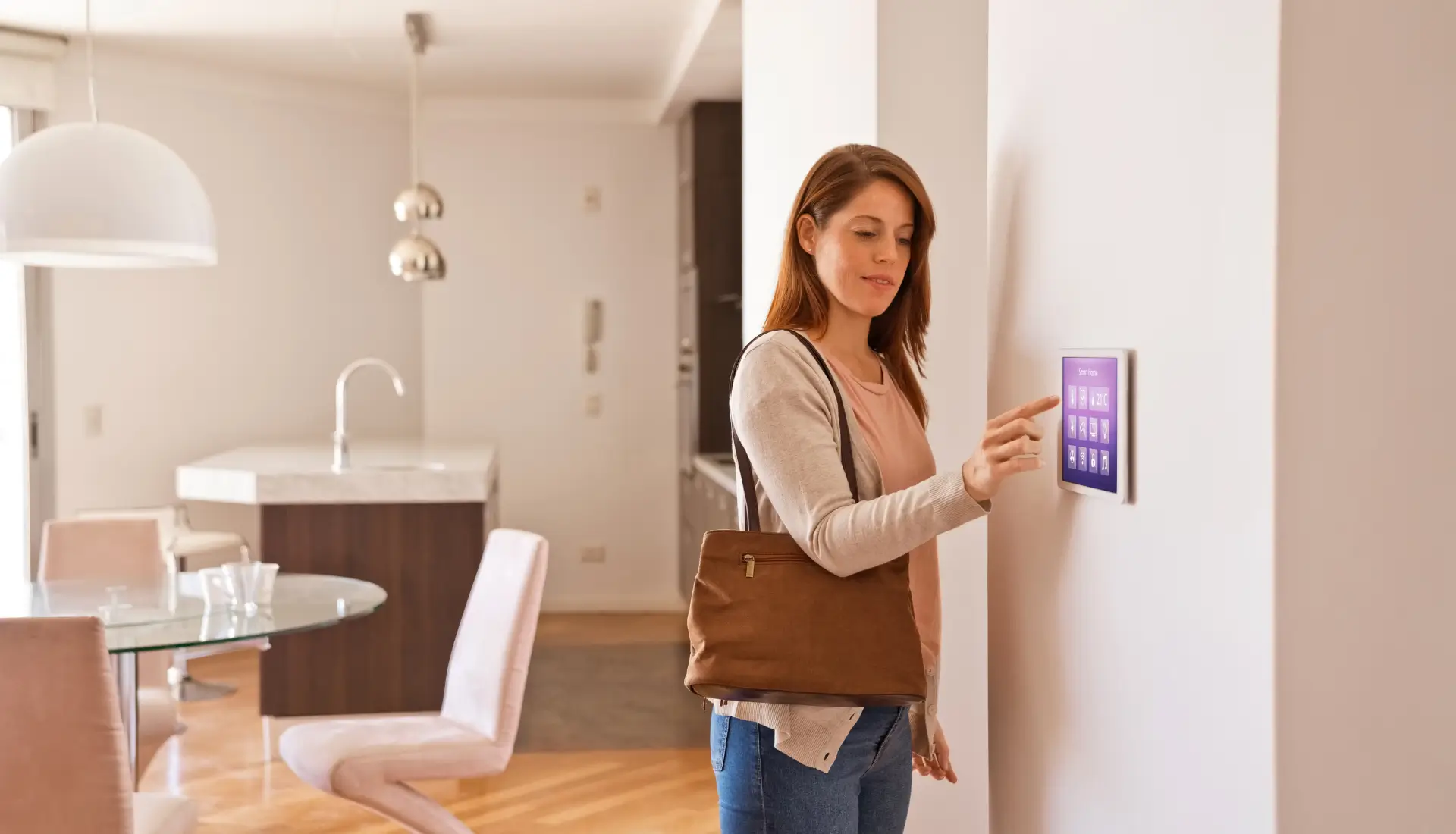A woman standing in a modern home interior, interacting with a smart home control panel mounted on the wall. The room features a dining area with light-colored furniture and wooden flooring.